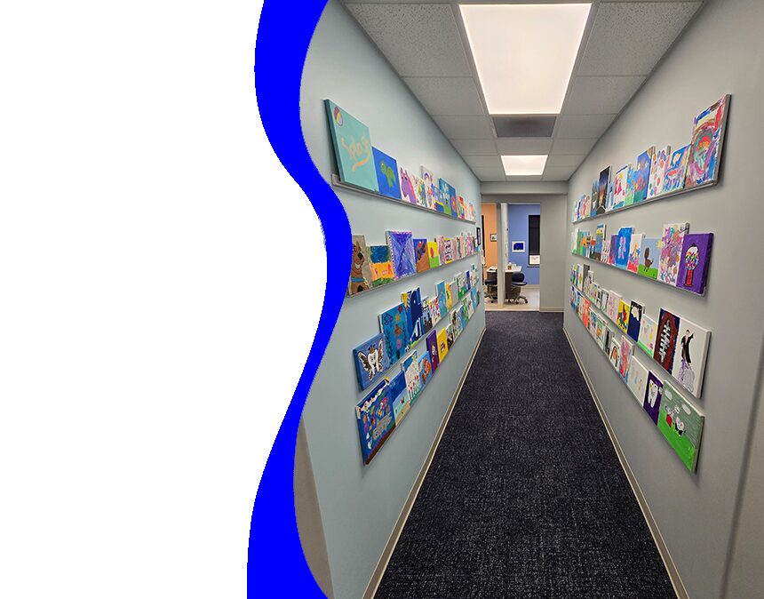 The image shows an interior view of a well-lit room with a blue and white color scheme. It appears to be a waiting area, possibly within a medical office, with shelves stocked with various items such as books, cards, and small gifts. The flooring is carpeted in neutral tones, and the walls are painted in a light hue. There s a large window on the right side of the room, allowing natural light to enter.