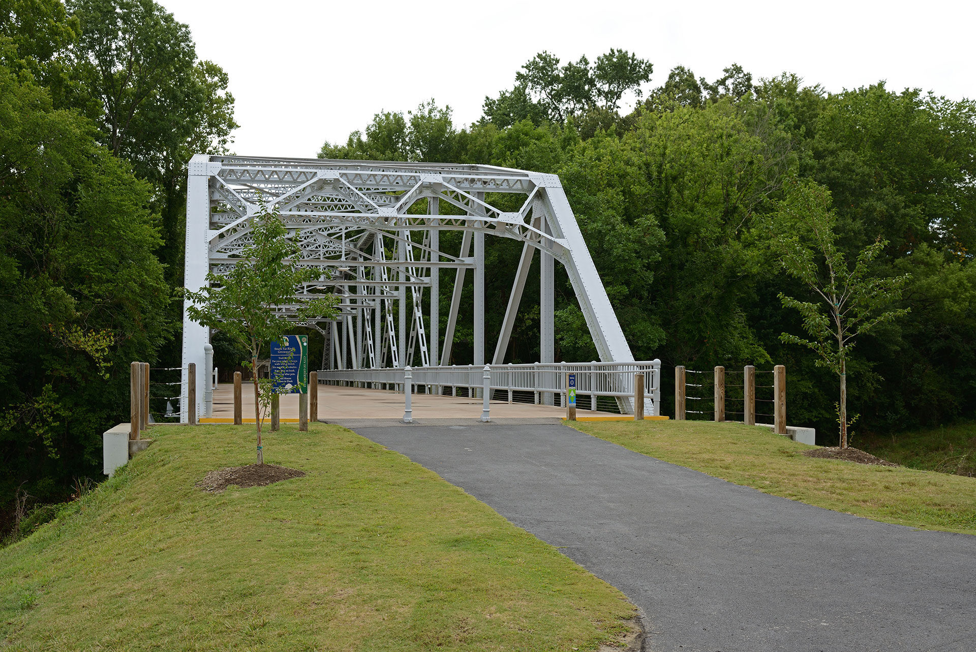 The image depicts a bridge overpassing a road next to a grassy area with trees and a clear sky.