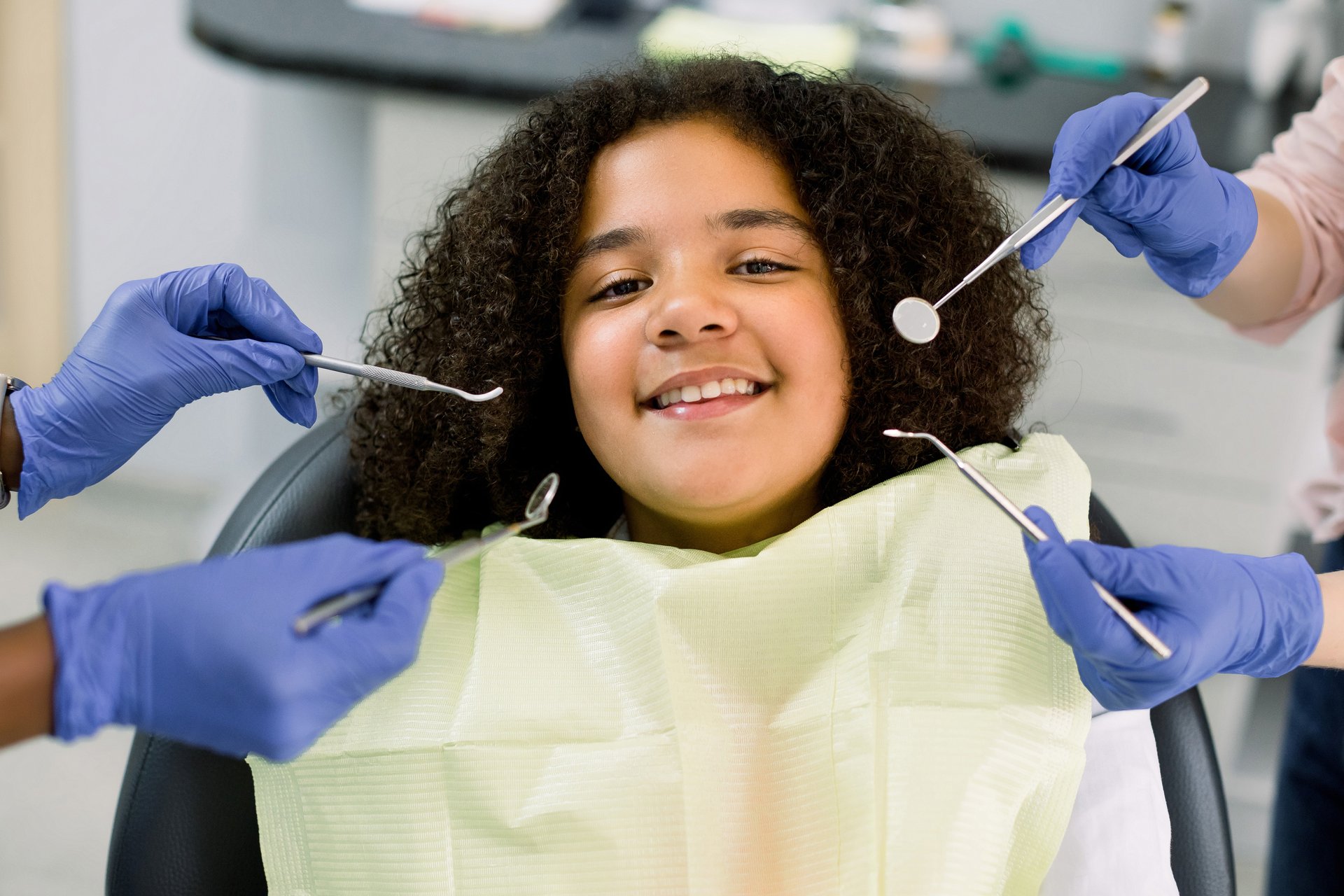 The image shows a young girl sitting in a dental chair, smiling at the camera while holding a teddy bear, with a woman behind her who appears to be a dental professional.